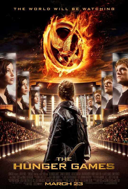Katniss se encuentra frente a carteles de ella y Peeta con el símbolo del Sinsajo en llamas en el centro en el cartel de la primera película de Los Juegos del Hambre.