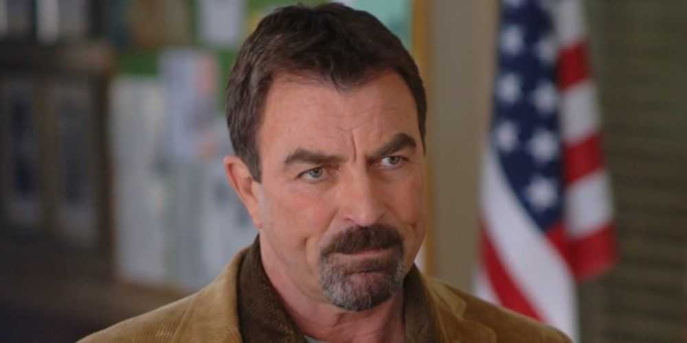 Tom Selleck con una bandera estadounidense detrás de él como Jesse Stone en Death in Paradise